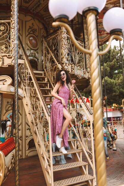 Jolie dame aux cheveux bouclés noirs en lunettes de soleil et robe debout avec des bonbons sucettes à la main et regardant joyeusement de côté tout en passant du temps sur un beau carrousel dans un parc d'attractions