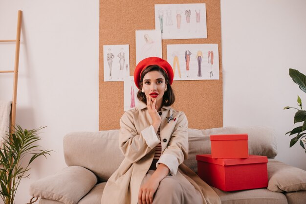 Jolie dame en béret rouge et trench beige assis sur un canapé. Fille élégante aux cheveux noirs en chapeau lumineux et boucles d'oreilles posant sur la caméra près des boîtes.