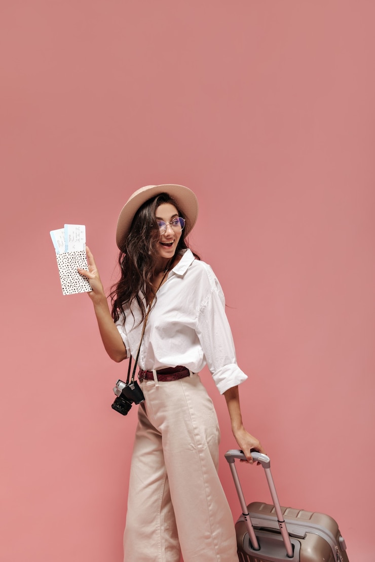 Photo gratuit jolie dame bouclée aux cheveux bruns en chemise blanche à manches larges, pantalon beige en ceinture moderne et lunettes élégantes posant avec des billets d'avion