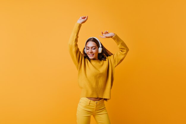 Jolie dame brune en pantalon jaune appréciant une bonne chanson. femme insouciante en pull doux dansant sur le mur orange.