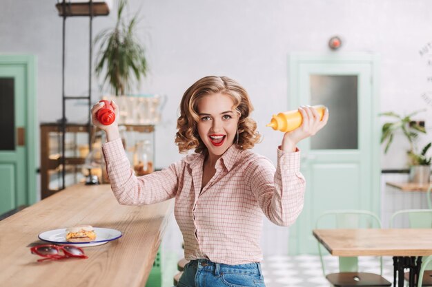 Jolie dame en chemise assise avec des bouteilles de ketchup et de moutarde dans les mains et regardant joyeusement à huis clos tout en passant du temps au café