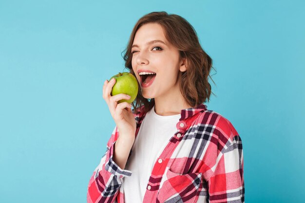 Jolie dame en chemise à carreaux mangeant une pomme verte et souriant joyeusement regardant à huis clos sur fond bleu coloré