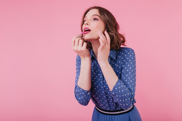 Jolie Dame Européenne Aux Yeux Sombres Faisant Du Maquillage Avec La Bouche Ouverte. Fille Frisée Agréable Dans Des Vêtements Bleus à L'aide De Rouge à Lèvres Rose.