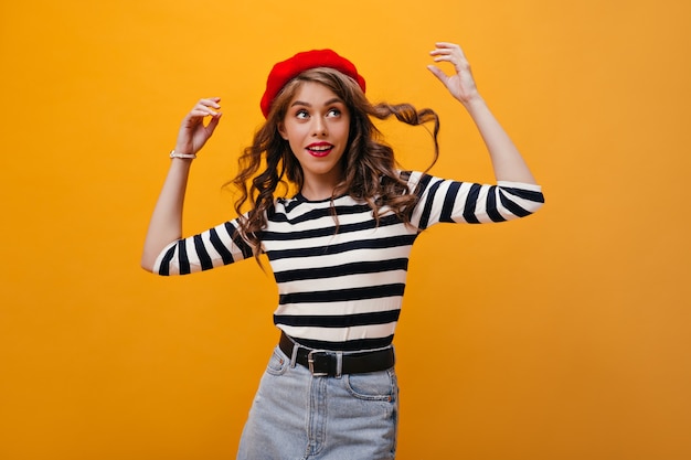 Jolie dame joue ses cheveux bouclés sur fond orange. Fanny jeune femme en béret rouge, chemisier rayé et jupe en jean posant sur fond isolé.