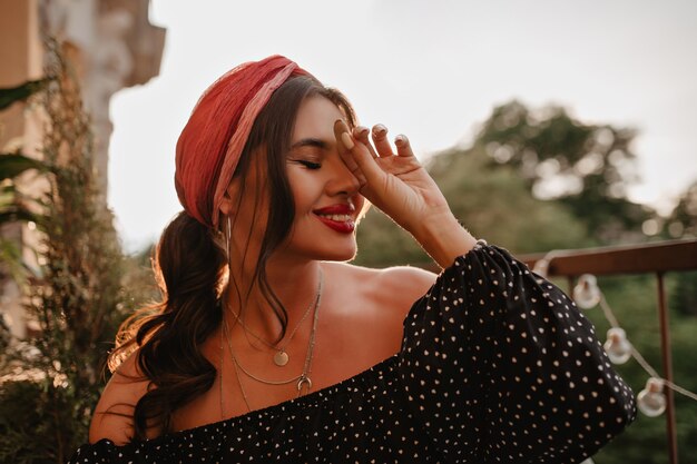 Jolie dame joyeuse aux cheveux noirs bouclés, accessoires en argent et bandana rose cool en chemise noire à manches longues souriant sur la terrasse.