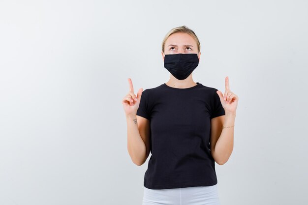 Jolie dame pointant vers le haut en t-shirt noir, masque noir et semblant reconnaissante isolée