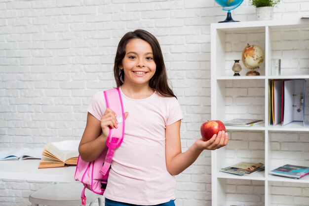 Jolie écolière debout avec sac à dos et pomme en salle de classe