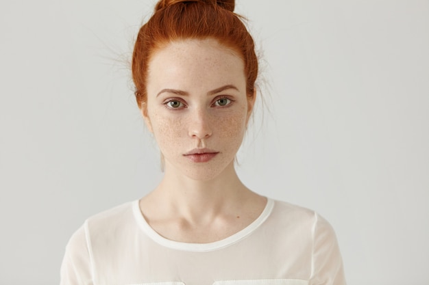 Jolie étudiante aux cheveux roux en noeud se détendre à la maison après le collège. Tête de tendre charmante jeune femme avec des taches de rousseur portant chemisier blanc posant