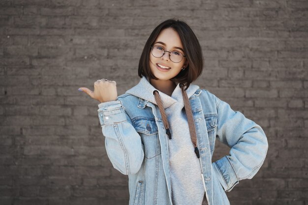 Jolie étudiante invitant les gens à rejoindre des cours de langue, recommander un centre d'éducation pour les jeunes, pointer le pouce vers la gauche et une caméra souriante, avoir l'air optimiste, recommander un endroit génial.