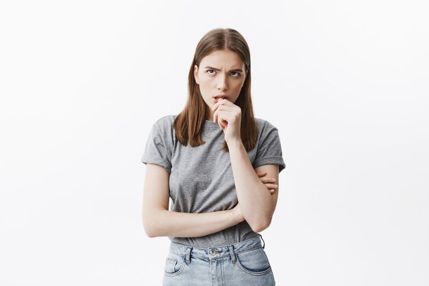 Une jolie étudiante malheureuse aux cheveux noirs en t-shirt gris ronge les doigts, regardant de côté avec un regard agité, ne peut pas se détendre en attendant les résultats du test à l'université.