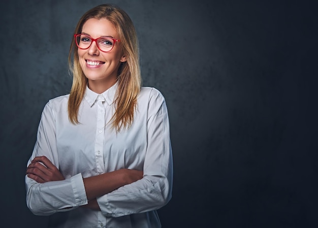 Jolie femme d'affaires blonde dans une chemise blanche, des lunettes et des bras croisés sur fond gris.