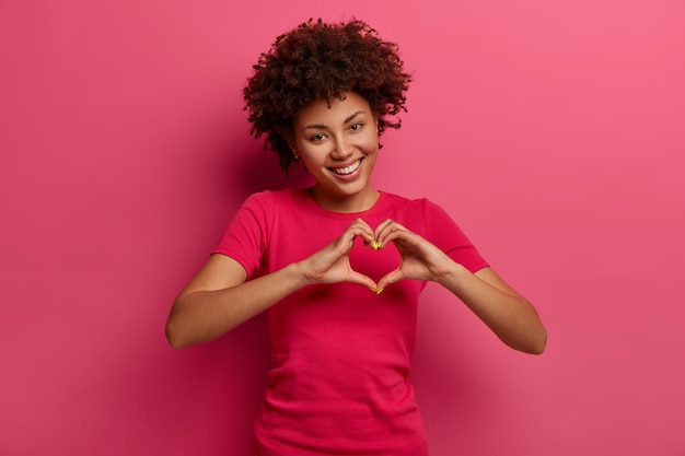 Jolie femme afro-américaine bouclée avoue amoureuse, fait un geste de cœur, montre ses vrais sentiments, a une expression heureuse, porte un t-shirt rouge décontracté, pose sur un mur rose. Concept de relation