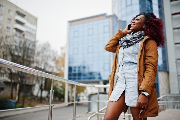 Jolie femme afro-américaine bouclée en manteau marron posée près des garde-corps contre un bâtiment moderne à plusieurs étages parlant au téléphone mobile