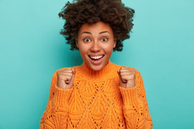 Une jolie femme afro-américaine charismatique applaudit la victoire, lève les poings fermés, célèbre quelque chose, sourit largement, montre des dents blanches, porte un pull orange tricoté