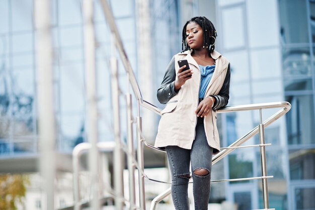 Jolie femme afro-américaine avec des dreads en veste posée près des garde-corps contre un bâtiment moderne à plusieurs étages regardant un téléphone mobile