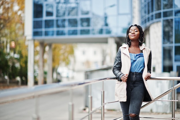 Jolie femme afro-américaine avec des dreads en veste posée près des garde-corps contre un bâtiment moderne à plusieurs étages