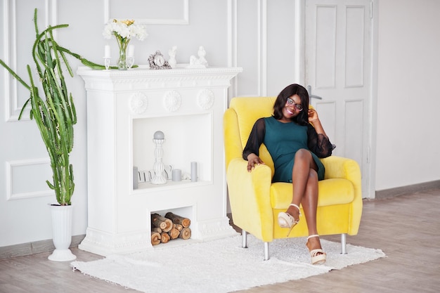 Jolie femme afro-américaine à lunettes posée dans la chambre assise sur une chaise jaune