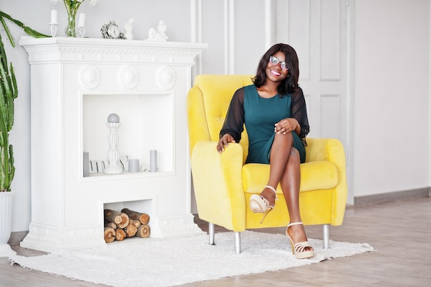 Jolie femme afro-américaine à lunettes posée dans la chambre assise sur une chaise jaune