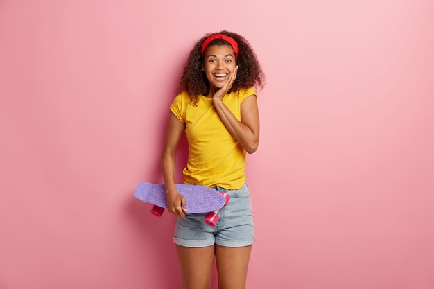 Une jolie femme afro satisfaite a une activité motrice, tient une planche à roulettes, se sent détendue et ravie