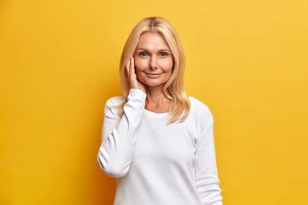 Jolie femme d'âge moyen tendre avec des cheveux blonds a une peau saine et ridée porte un maquillage minimal habillé en cavalier blanc occasionnel pose à l'intérieur