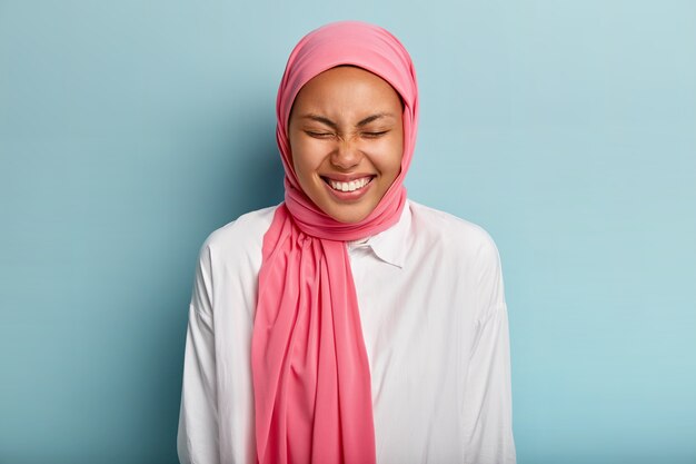Jolie femme arabe rit avec un large sourire, garde les yeux fermés, exprime de bonnes émotions