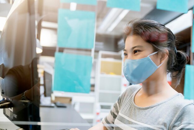 Jolie femme asiatique bleu marine pull travaillant tout en portant un masque de protection contre les virus travaillant dans un espace de co-woking qui a une cloison en plastique distanciation sociale pour le siège nouveau mode de vie normal