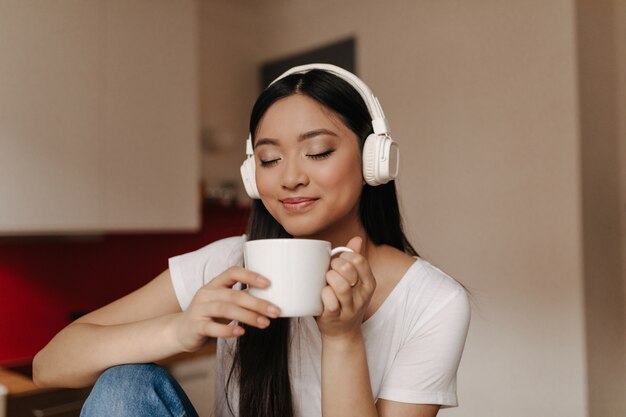 Jolie femme asiatique dans les écouteurs inhaler l'arôme du thé, tenant la tasse et souriant