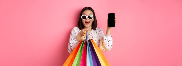 Photo gratuite jolie femme asiatique montrant une application pour smartphone et des sacs à provisions achetant en ligne via l'application standi