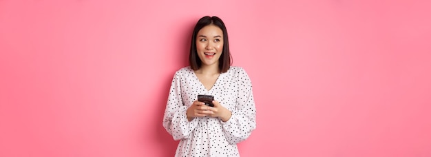 Photo gratuite jolie femme asiatique pensant et souriant regardant à gauche rêveuse tout en envoyant des messages sur un smartphone en naviguant en ligne