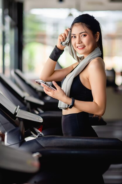 Une jolie femme asiatique portant des vêtements de sport et une montre intelligente se repose sur un tapis roulant, utilise un smartphone et une application d'entraînement pour montre intelligente et écoute de la musique dans une salle de sport moderne