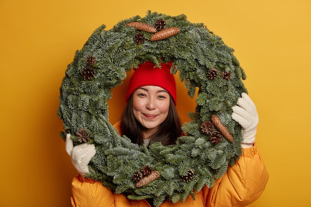 Jolie femme asiatique porte des couvre-chefs et des gants chauds, regarde à travers une couronne de Noël traditionnelle faite à la main, étant de bonne humeur festive, anticipe les vacances d'hiver, isolé sur le mur jaune du studio
