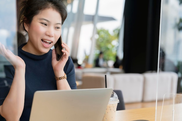 Jolie femme asiatique pull bleu marine communication à la main avec un smartphone travaillant dans un espace de co-woking qui a une cloison en plastique distanciation sociale pour le siège nouveau mode de vie normal