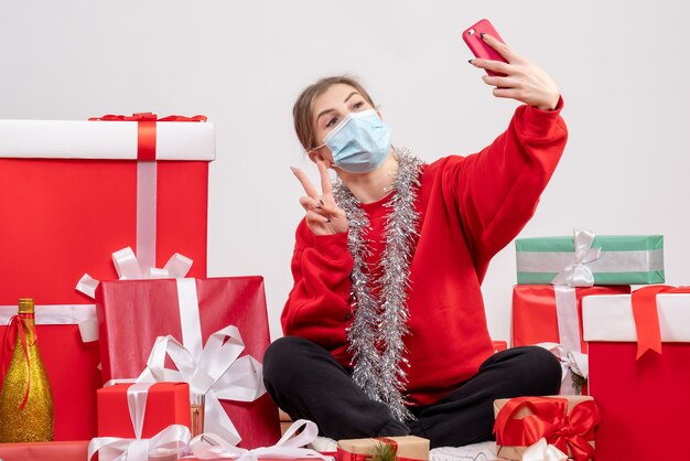 Jolie femme assise autour de cadeaux de Noël prenant selfie sur blanc