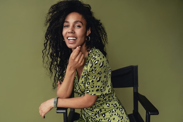 Jolie femme assise sur une chaise dans une élégante robe imprimée léopard verte isolée sur un mur de studio vert