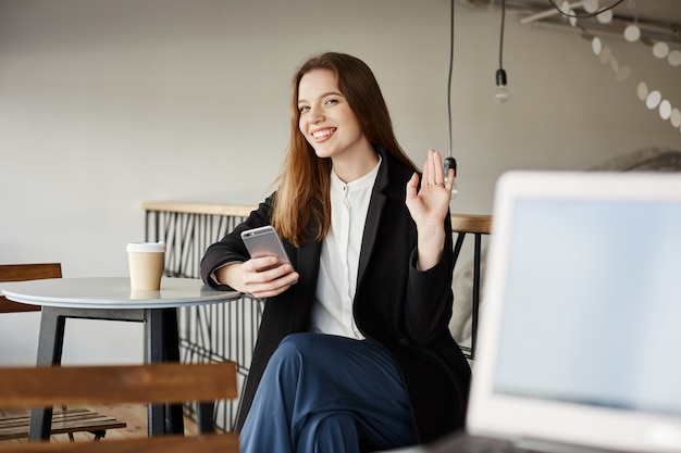 Jolie femme au café avec smartphone, agitant la main pour saluer, dire bonjour