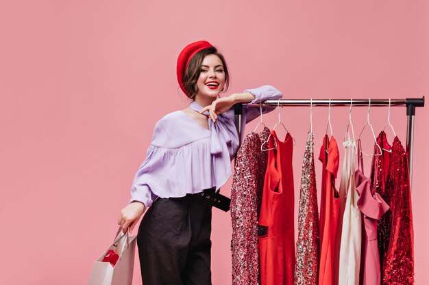 Jolie femme au chapeau lumineux et chemisier violet se penche sur le stand avec des robes et posant avec le paquet sur fond isolé.