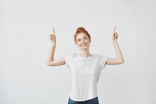 Jolie femme au gingembre avec des taches de rousseur souriant pointant les doigts vers le haut.