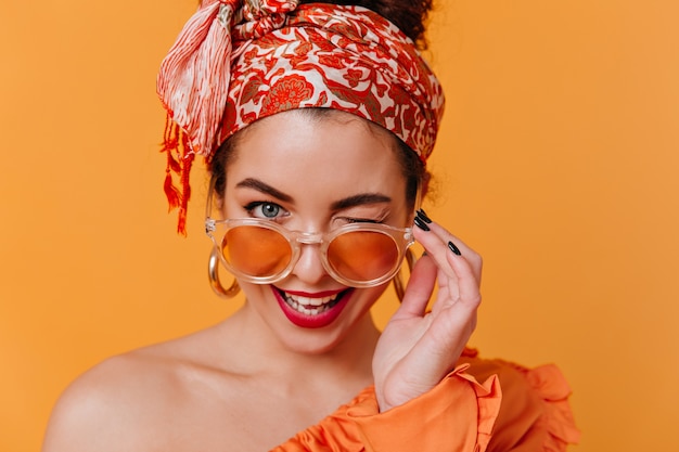 Photo gratuite jolie femme aux boucles d'oreilles massives et serre-tête de style africain enlève ses lunettes orange et fait un clin d'œil coquette.