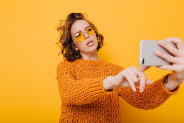 Jolie femme aux cheveux bouclés courts tenant le smartphone et en tapant un message devant un mur jaune