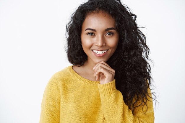 Jolie femme aux cheveux bouclés en pull jaune souriante ravie, comme ce qu'elle considère comme un bon choix, touche le menton réfléchi, réfléchit à une bonne idée