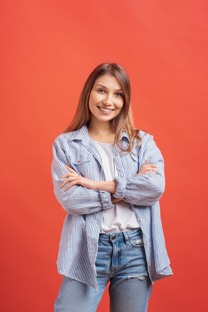 Jolie femme aux cheveux longs posant avec les bras croisés, expression du visage souriant sur le mur rouge.