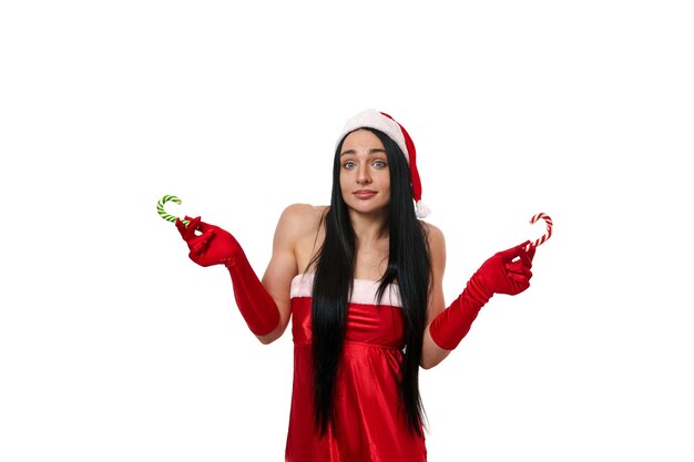 Jolie femme aux cheveux noirs sexy en costume de carnaval du père noël tenant des sucettes rayées vertes rouges et blanches, des cannes de bonbon de noël et regardant la caméra isolée sur fond blanc. copier l'espace publicitaire
