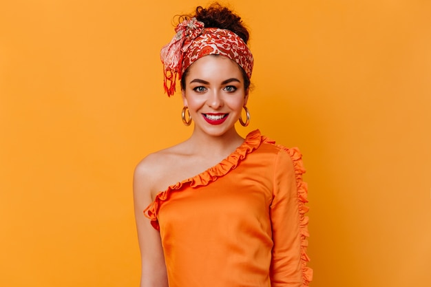 Jolie femme aux lèvres rouges en foulard et chemisier en soie avec sourire se penche sur la caméra sur l'espace orange.