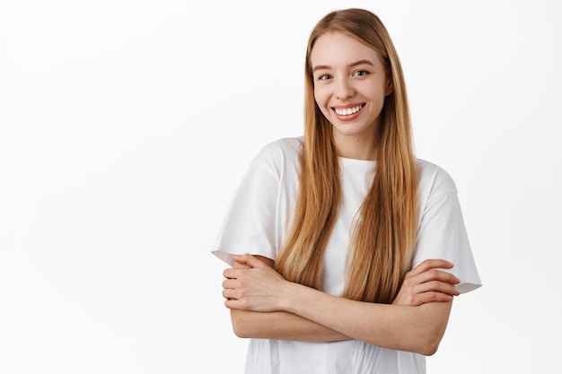 Jolie femme aux longs cheveux blonds raides, les bras croisés sur la poitrine, l'air confiante et déterminée, souriante heureuse, debout en t-shirt contre le mur blanc