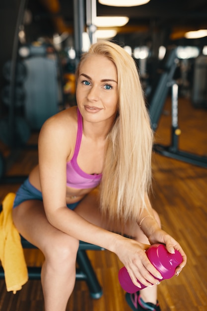 Photo gratuite jolie femme aux longs cheveux blonds se détendre dans la salle de gym avec une bouteille d'eau rose