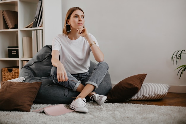 Jolie femme avec de belles boucles d'oreilles massives et en T-shirt blanc avec sourire, posant assis dans un fauteuil.