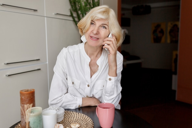 Jolie femme blonde d'âge moyen se détendre à la maison dans la cuisine et parler par téléphone mobile