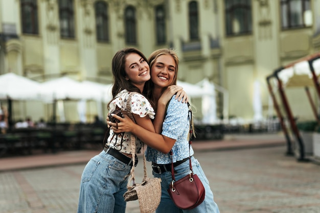 Jolie femme blonde en blouse bleue et fille en haut floral blanc câlin à l'extérieur Charmante dame brune en pantalon denim sourit et pose avec un ami à l'extérieur