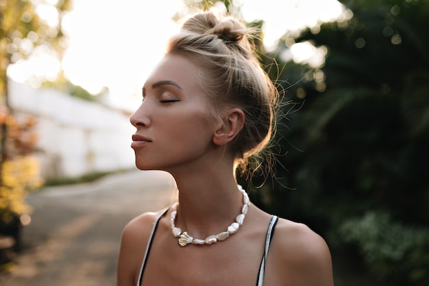 Photo gratuite jolie femme blonde bronzée avec chignon et collier de perles pose avec les yeux fermés à l'extérieur