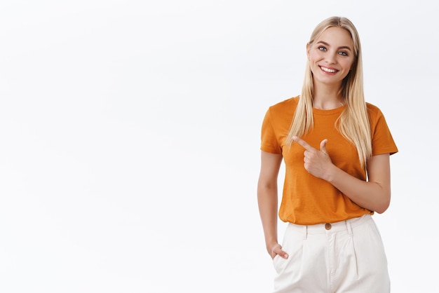 Jolie femme blonde caucasienne élégante et confiante en pantalon t-shirt orange tenir la main dans la poche pointant le doigt côté gauche fond souriant joyeusement suggérer de regarder fond blanc